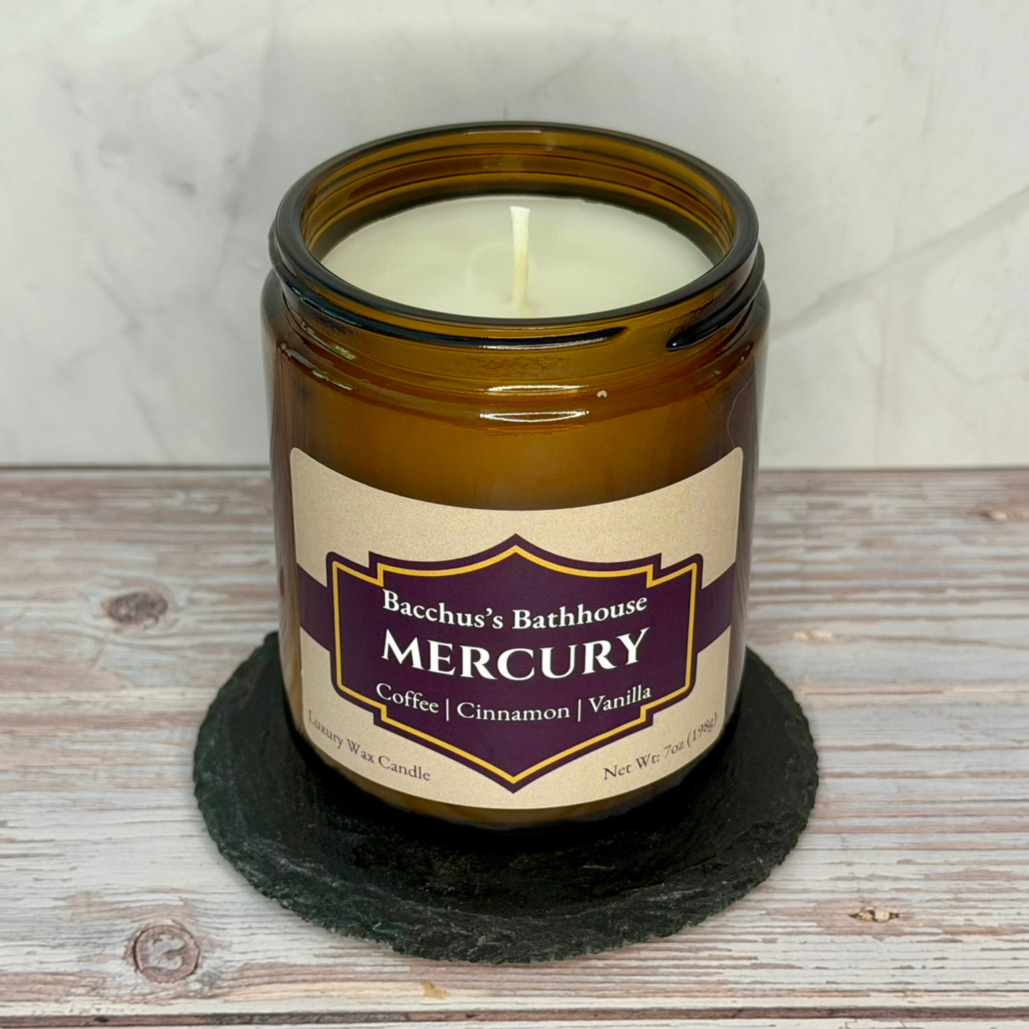 An amber glass jar candle with a cream-colored wax and label featuring a purple and gold design. The label reads 'Bacchus's Bathhouse MERCURY Coffee | Cinnamon | Vanilla'.