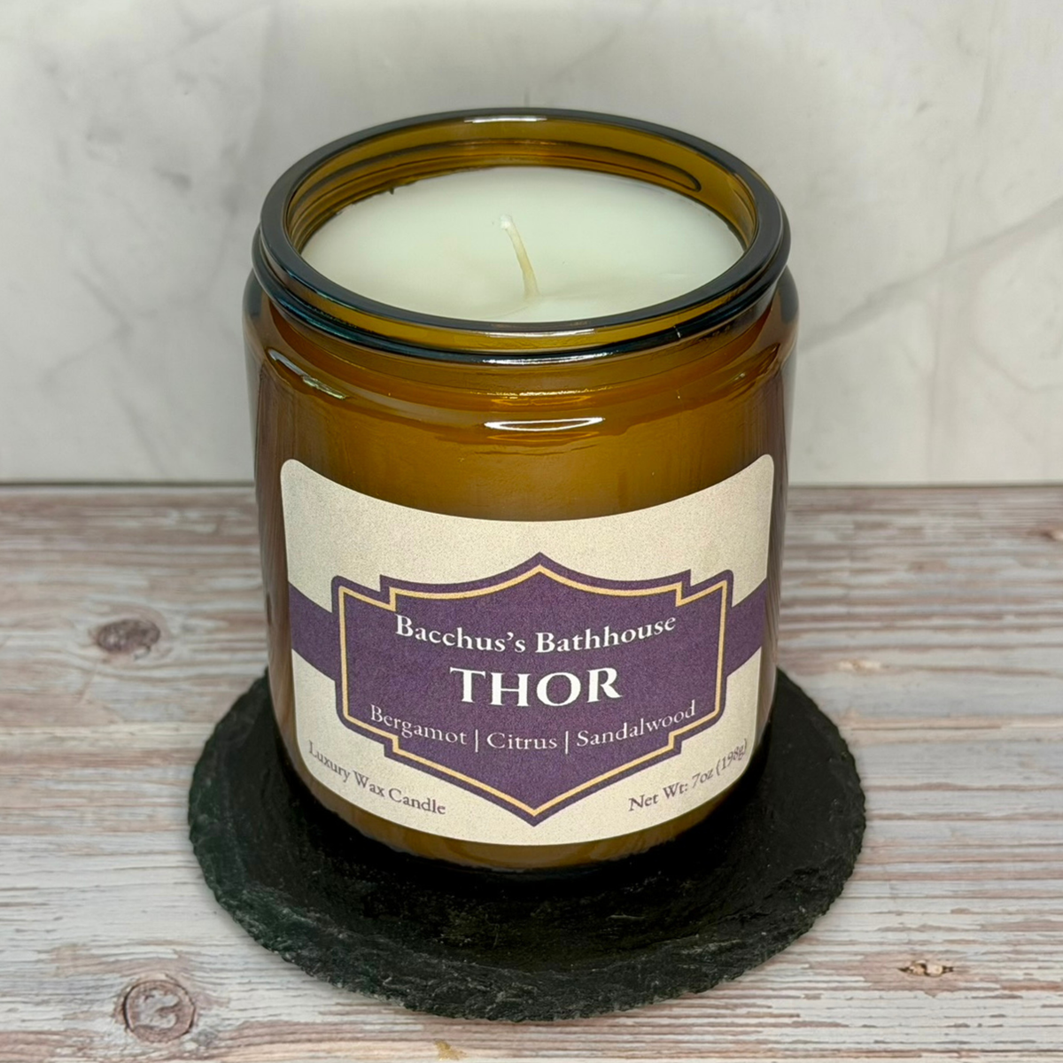 An amber glass jar candle with white wax sits on a dark round coaster on a light wood surface. The label reads 'Bacchus's Bathhouse THOR Bergamot | Citrus | Sandalwood'.
