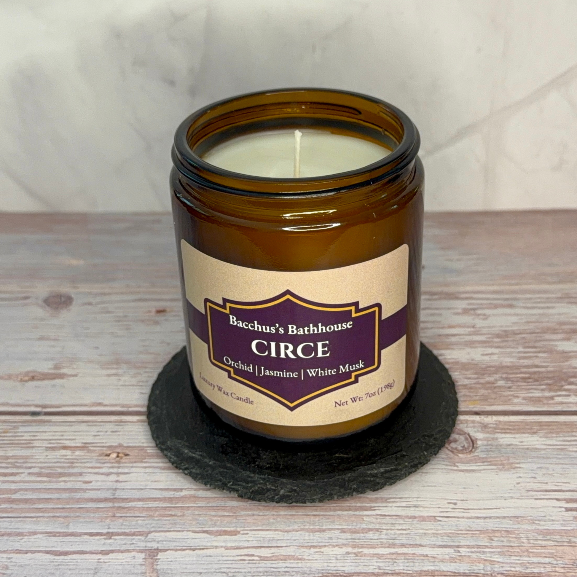 An amber glass jar candle with a cream label reading 'Bacchus's Bathhouse CIRCE' sits on a round black slate coaster over a rustic whitewashed wood surface.