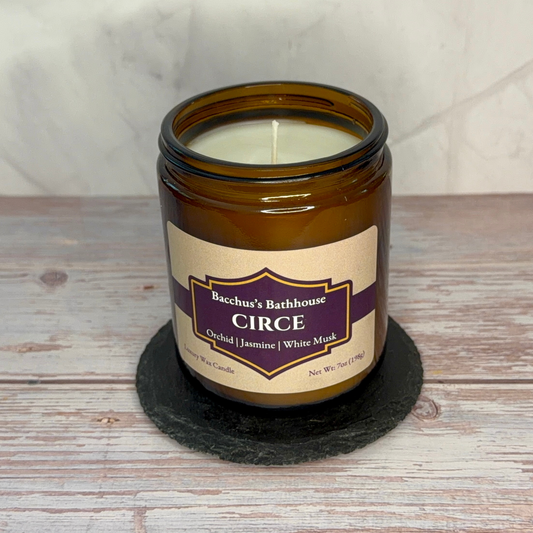An amber glass jar candle with a cream label reading 'Bacchus's Bathhouse CIRCE' sits on a round black slate coaster over a rustic whitewashed wood surface.