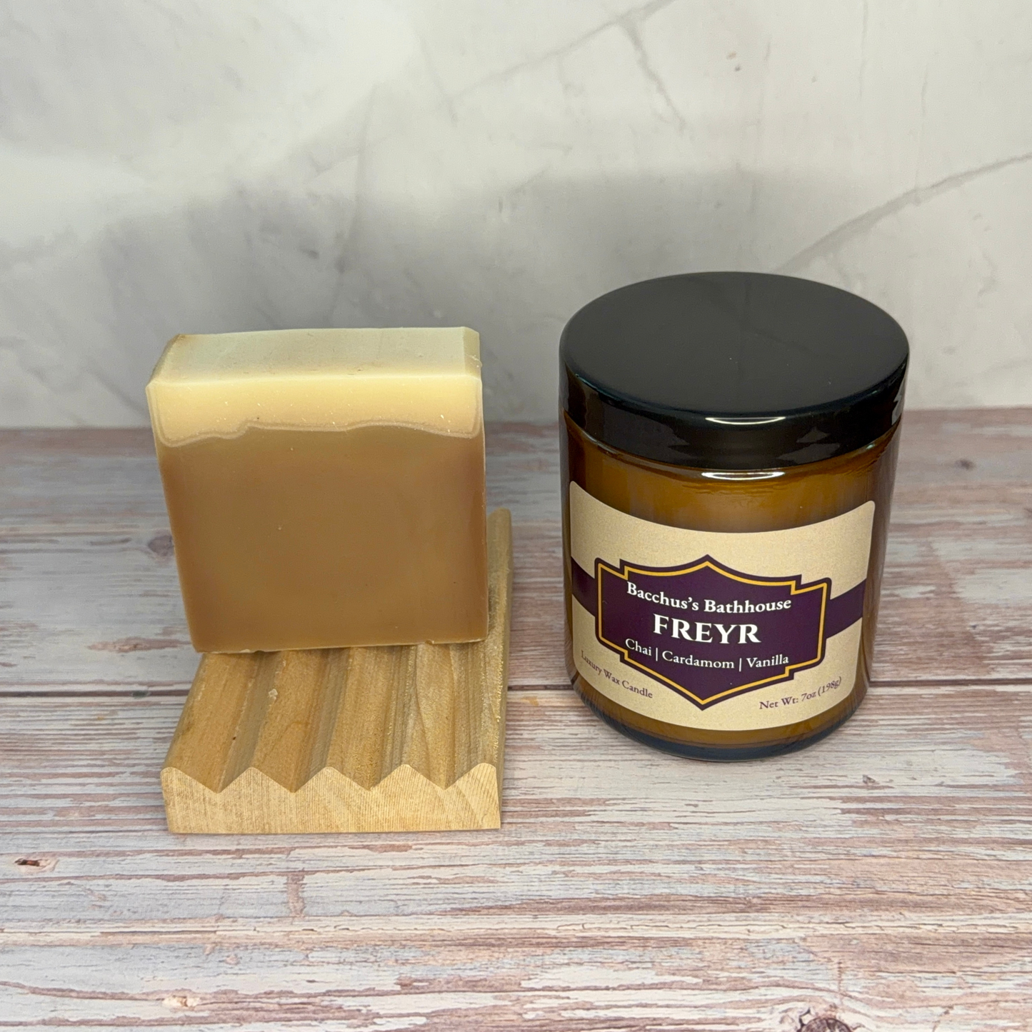 A handmade bar of soap in a light tan shade stands on a small wooden stand next to a candle jar labeled 'Bacchus's Bathhouse FREYR' on a rustic whitewashed wood surface.