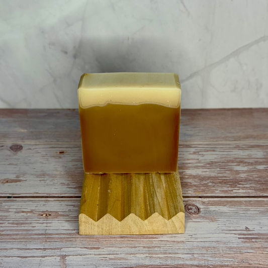 A rectangular bar of layered soap in amber and pale cream, displayed on a small wooden stand over a rustic wood surface with a light stone wall background.