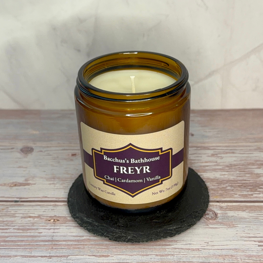 An amber glass jar candle labeled 'Bachus's Bathhouse FREYR' sits on a round black slate coaster over a whitewashed wooden surface, with a light stone wall behind it.