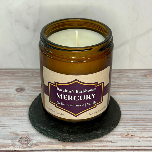An amber glass jar candle with a cream-colored wax and label featuring a purple and gold design. The label reads 'Bacchus's Bathhouse MERCURY Coffee | Cinnamon | Vanilla'.
