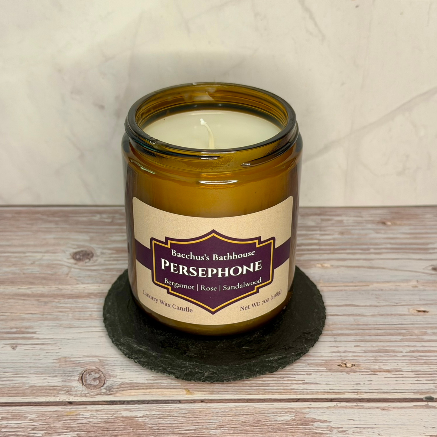 An amber glass jar candle labeled Bacchus's Bathhouse Persephone, placed on a round black coaster over a rustic wood surface against a light textured wall.