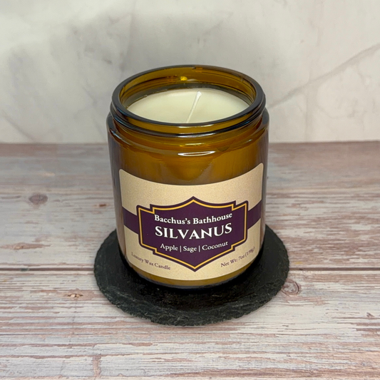An amber glass jar candle with a cream-colored wax and a beige label reading 'Bacchu's Bathhouse SILVANUS Apple | Sage | Coconut', sitting on a black coaster over a rustic wood surface.