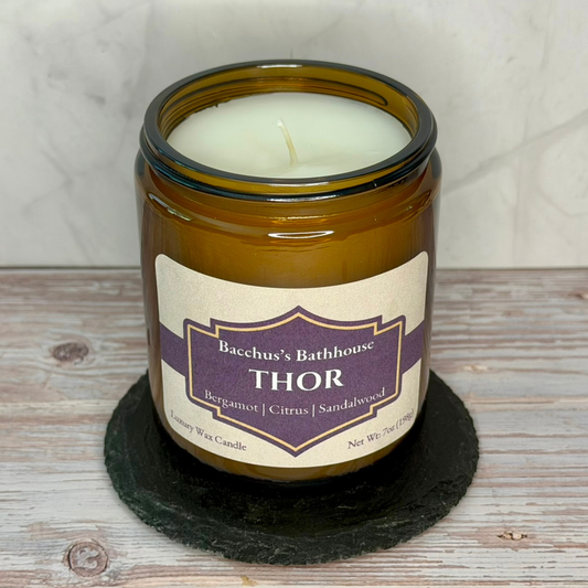An amber glass jar candle with white wax sits on a dark round coaster on a light wood surface. The label reads 'Bacchus's Bathhouse THOR Bergamot | Citrus | Sandalwood'.